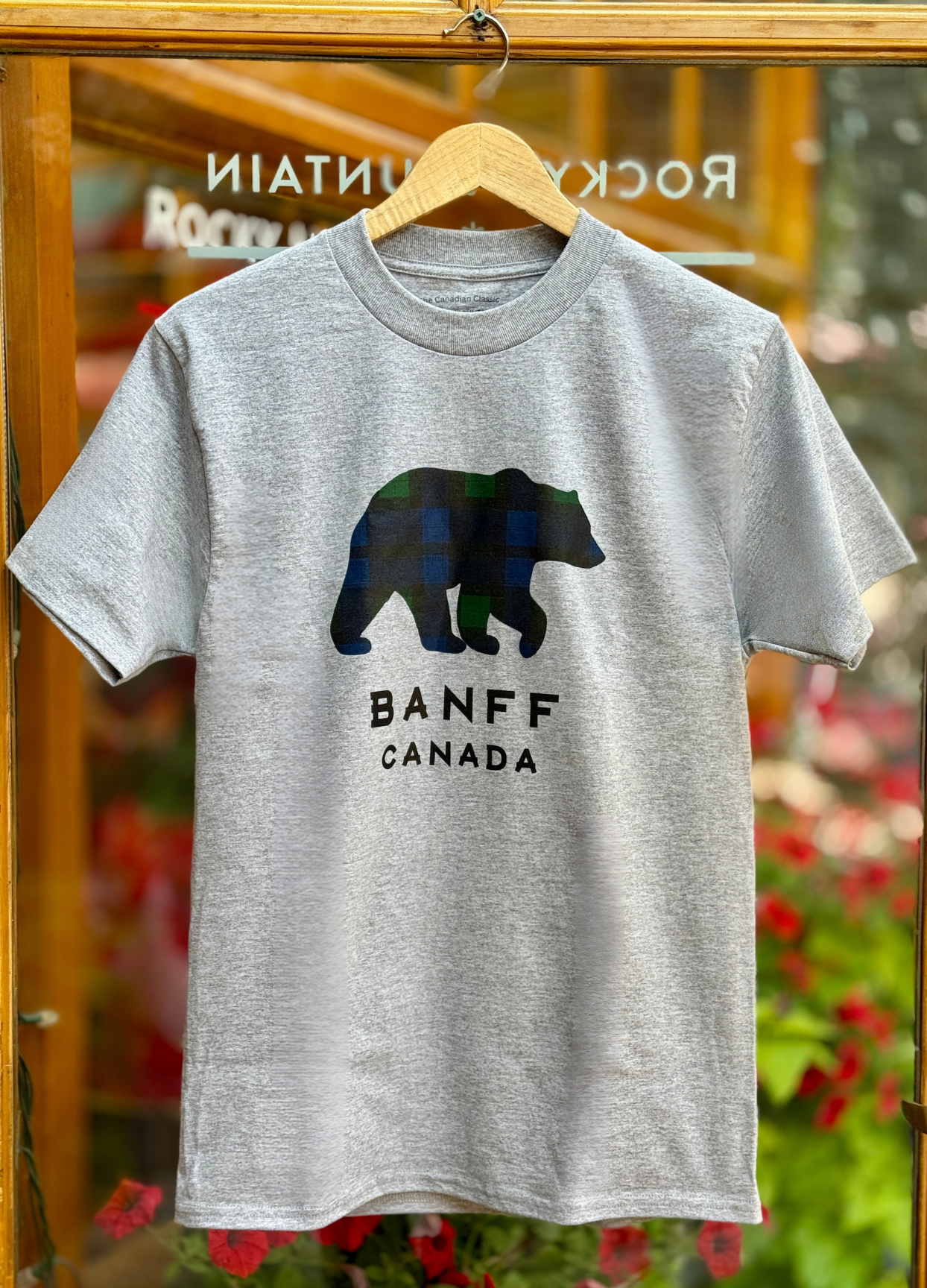 Grey t-shirt with a bear graphic and 'Banff Canada' text on a hanger against a blurred background.