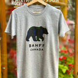 Gray t-shirt with a bear graphic and 'Banff Canada' text on a hanger in front of a store window.