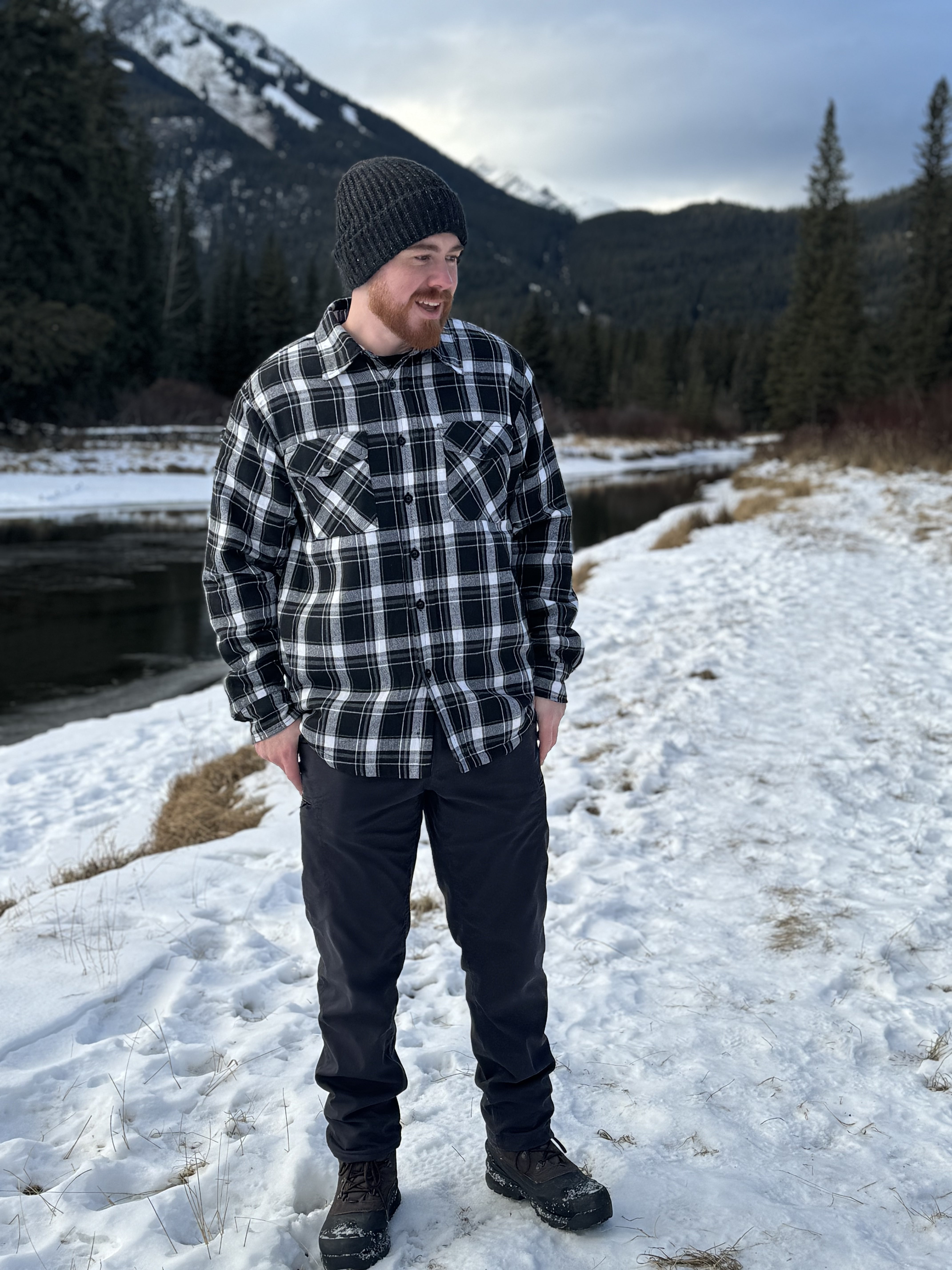 A man wearing a black and white plaid padded flannel jacket, standing near a snowy landscape.