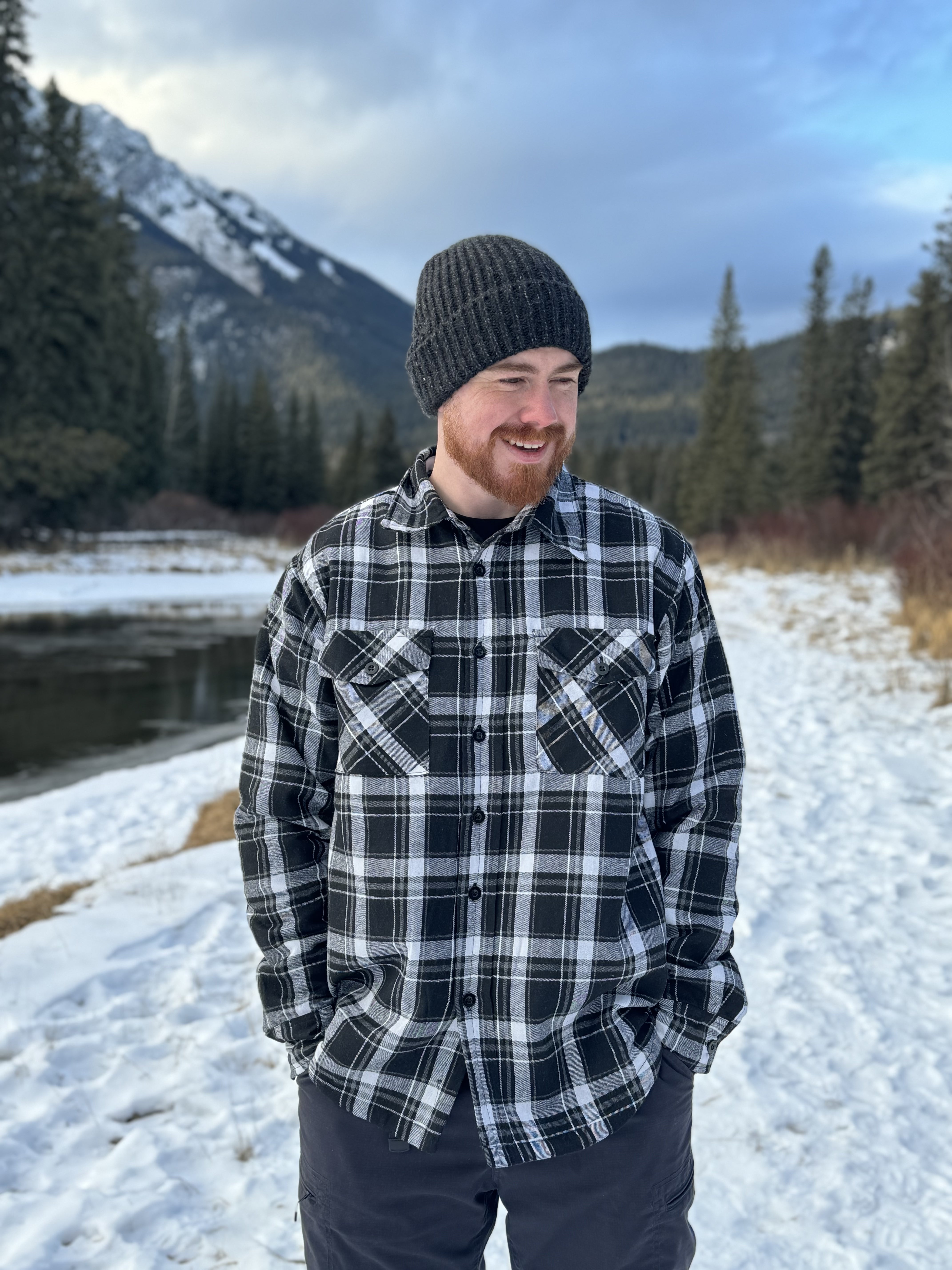 A man wearing a black and white plaid padded flannel jacket, standing near a snowy landscape.