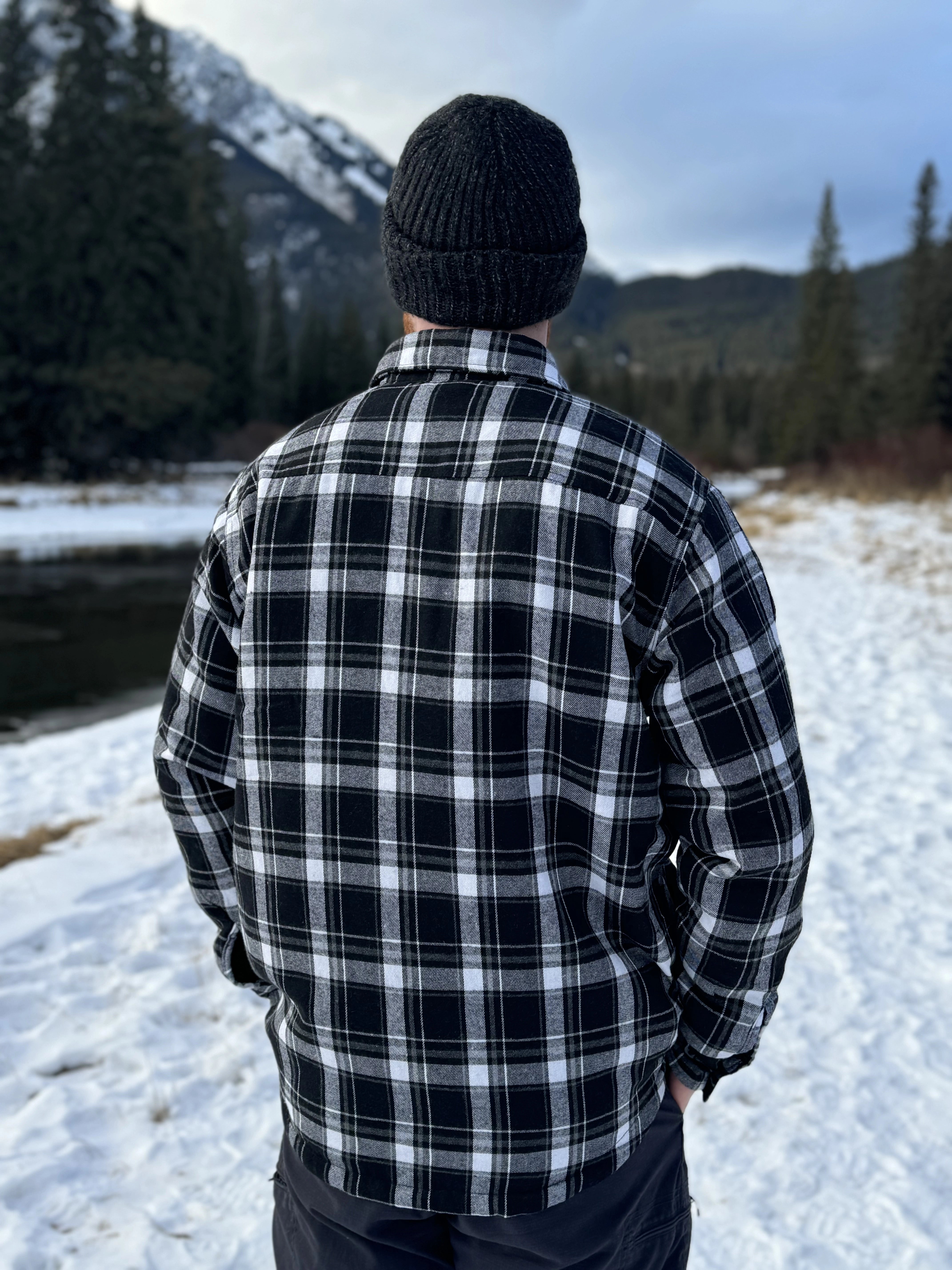 Back view of a man wearing a black and white plaid padded flannel jacket, standing near a snowy landscape.