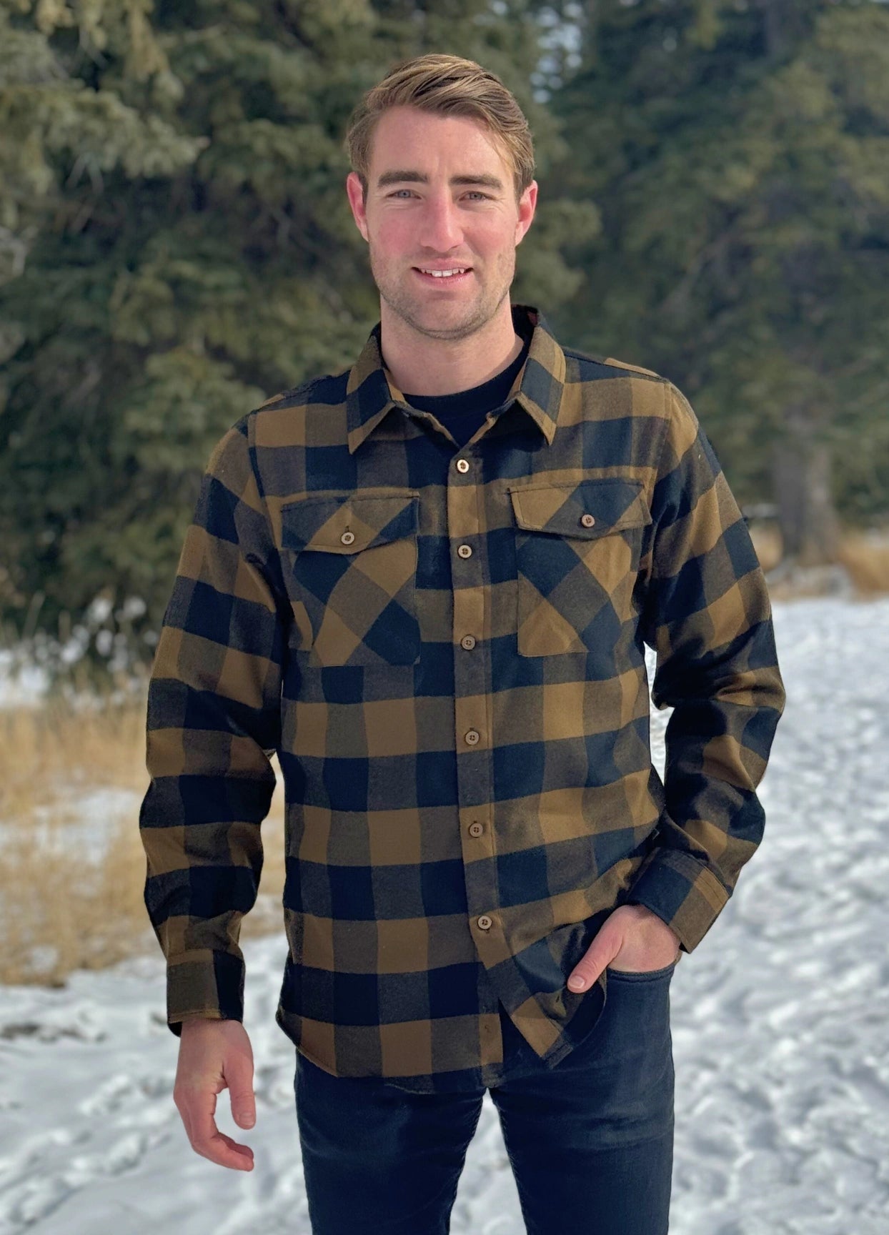 A man wearing a black and brown buffalo check flannel shirt with two flap breast pockets and two button cuff, standing with one hand in his front pocket.
