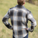 Back view of a person standing in an open field wearing a plaid flannel shacket with a herringbone weave, button-up collar, and long sleeves.