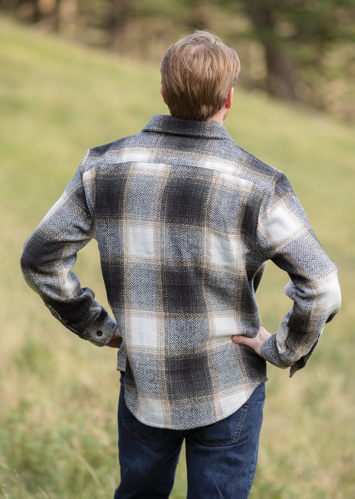 Back view of a person standing in an open field wearing a plaid flannel shacket with a herringbone weave, button-up collar, and long sleeves.