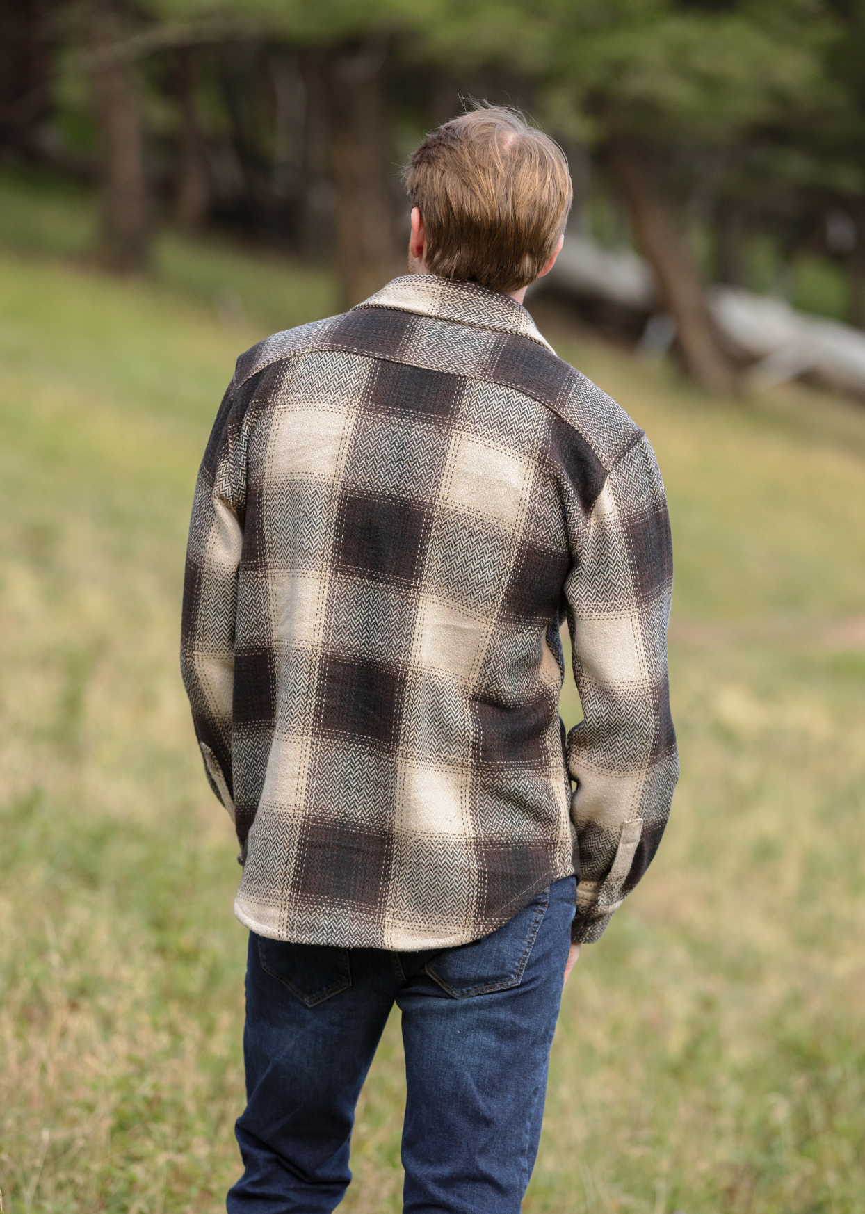 Back view of a person wearing a brown wool blend flannel jacket with herringbone weave, paired with blue jeans, standing in a natural outdoor setting.