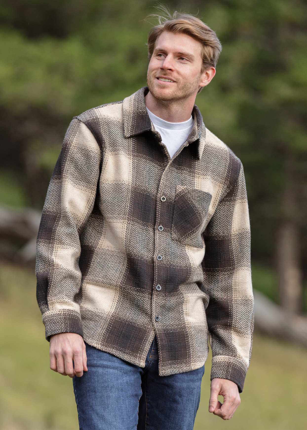 A person wearing a brown wool blend flannel jacket with herringbone weave, paired with blue jeans, standing in a natural outdoor setting.