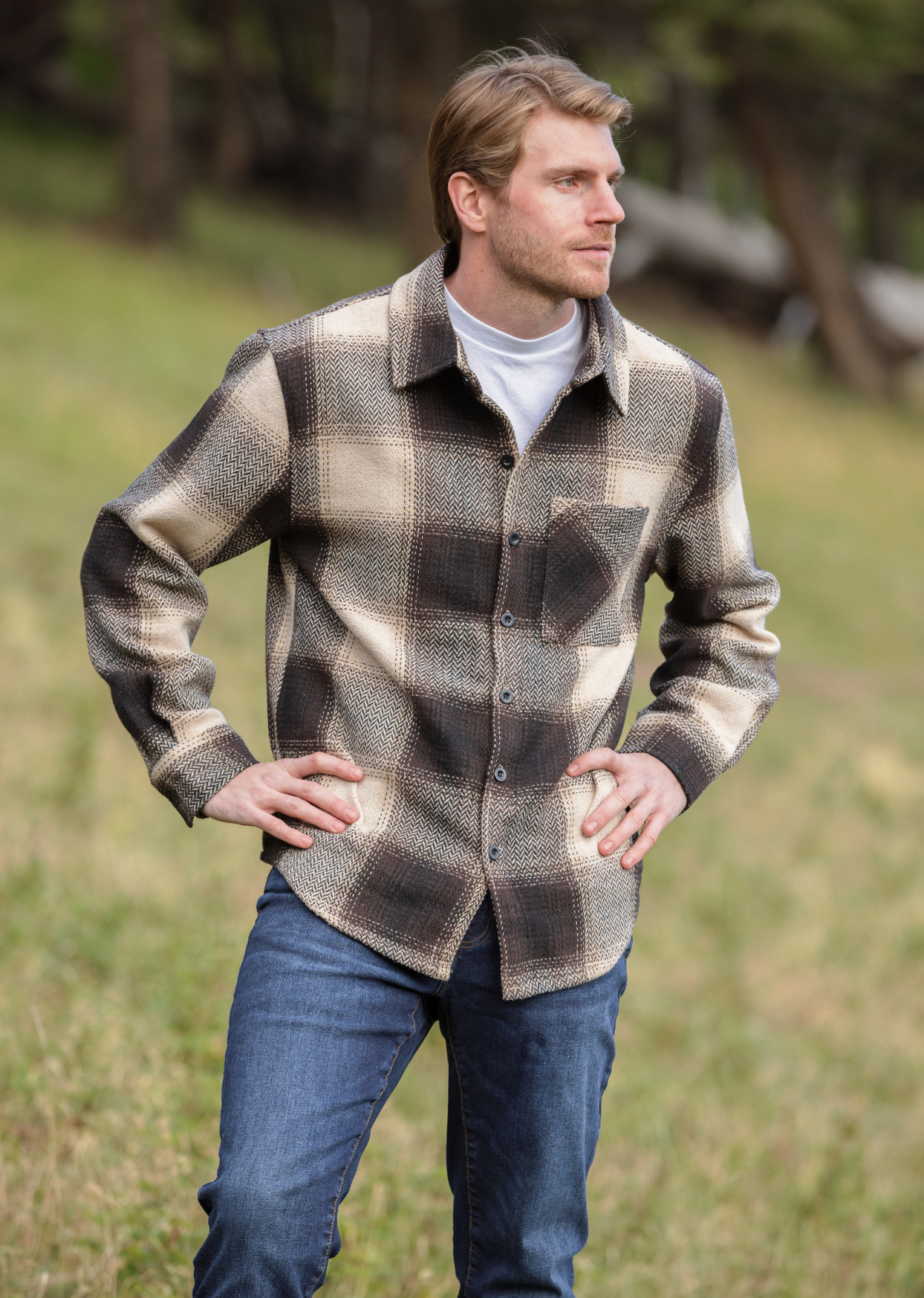 A person wearing a brown wool blend flannel jacket with herringbone weave, paired with blue jeans, standing in a natural outdoor setting.