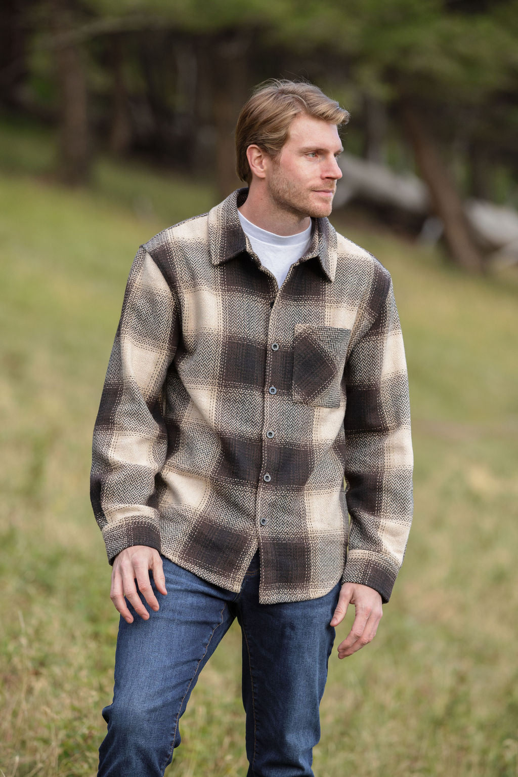 A person wearing a brown wool blend flannel jacket with herringbone weave, paired with blue jeans, standing in a natural outdoor setting.