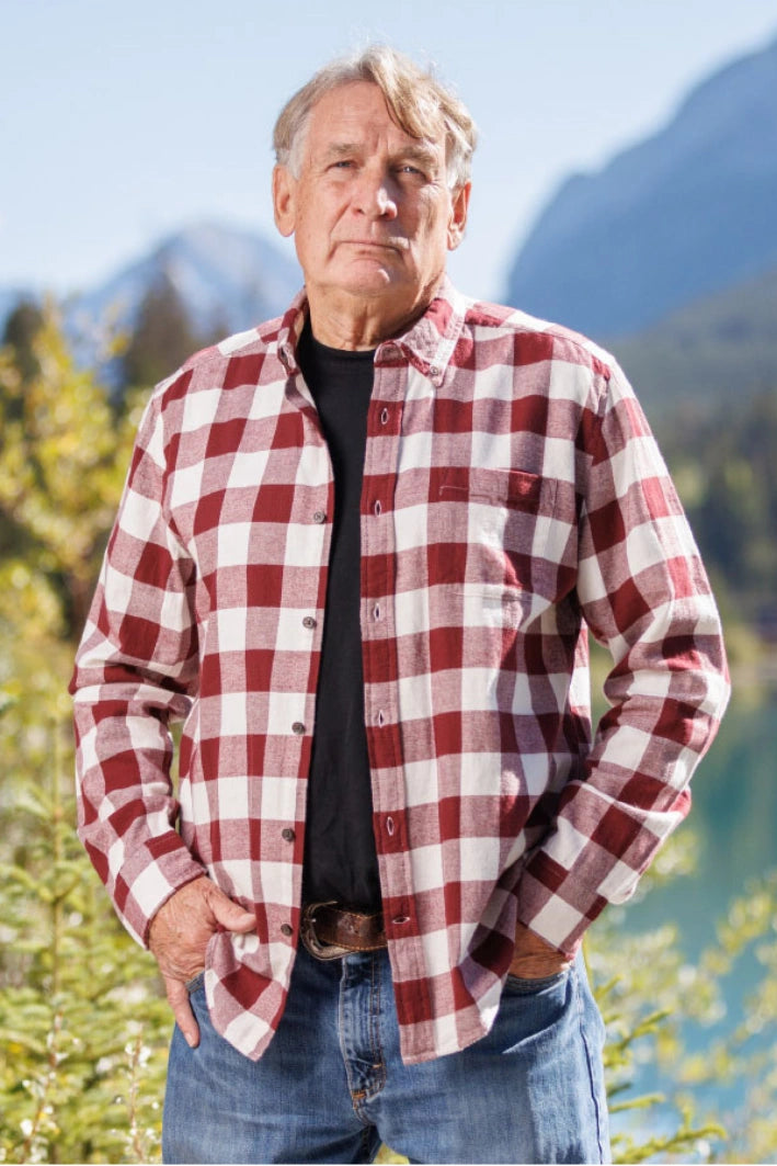 An older man with short grey hair wearing a burgundy and white buffalo check flannel shirt and blue jeans, standing near a body of water.