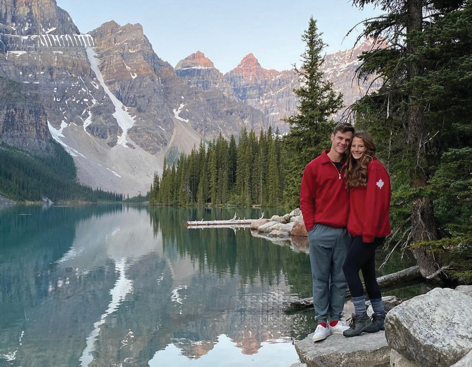 A couple standing in front of Moraine Lake in Banff National Park. Wearing our red canadian jackets with maple leaf patches on the arm. These jackets are made in canada.