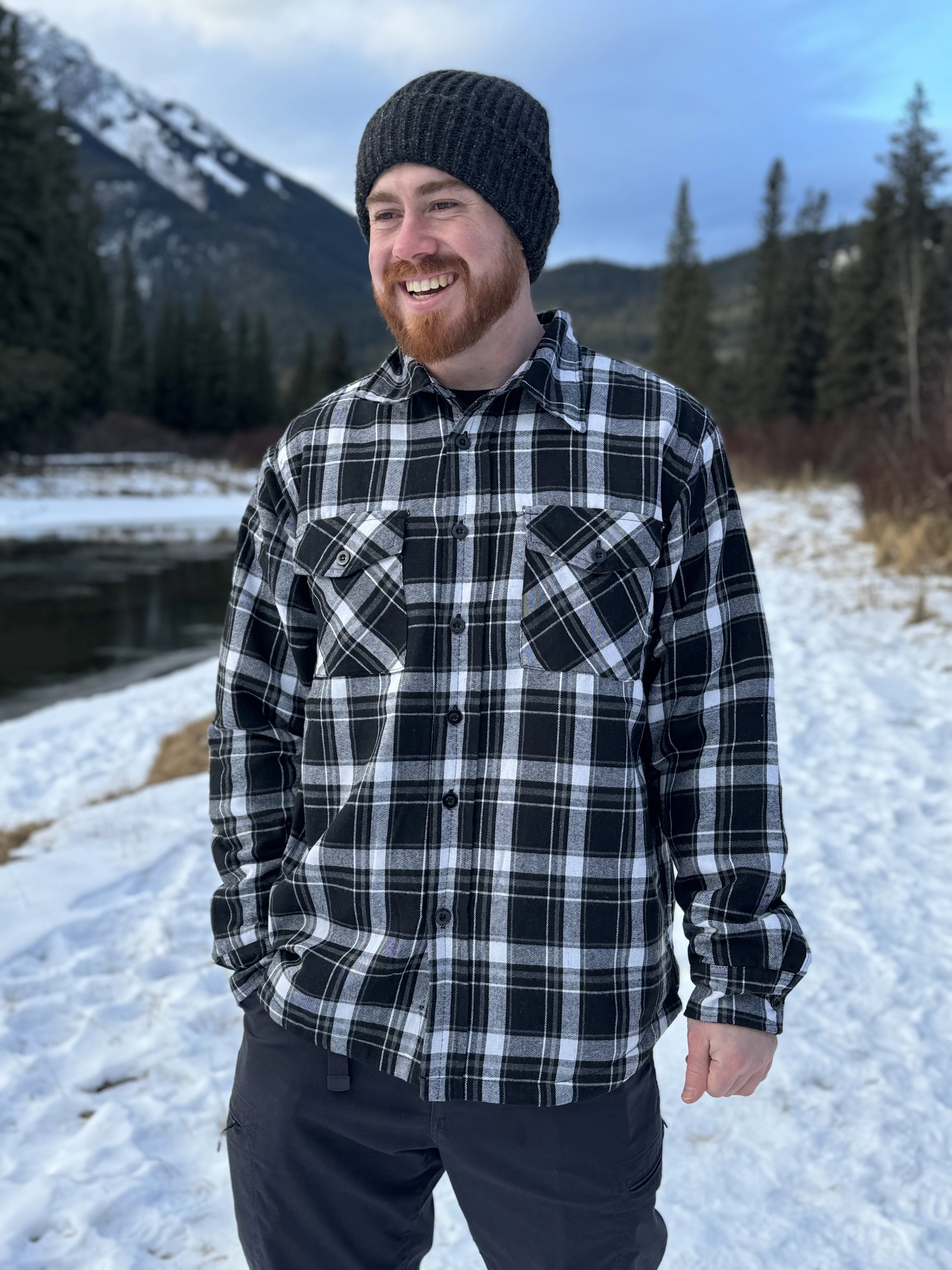 A man wearing a black and white plaid padded flannel jacket, standing near a snowy landscape.