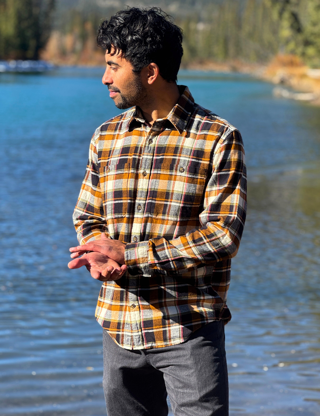 Man wearing a plaid flannel shirt standing by a lake with mountains in the background