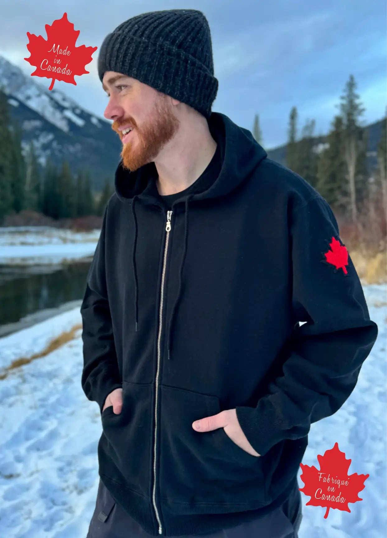 A person wearing a full zip black hoody with a kangaroo pocket, standing outdoors with snow and trees in the background.