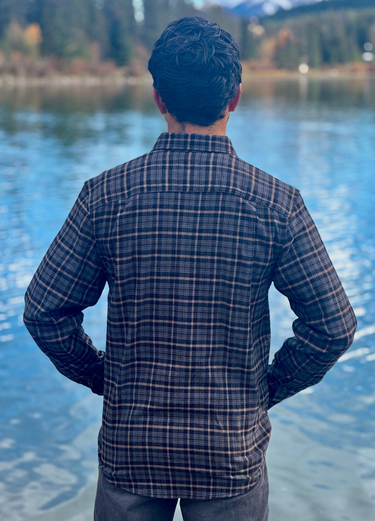 Person wearing a plaid shirt standing by a lake with mountains in the background