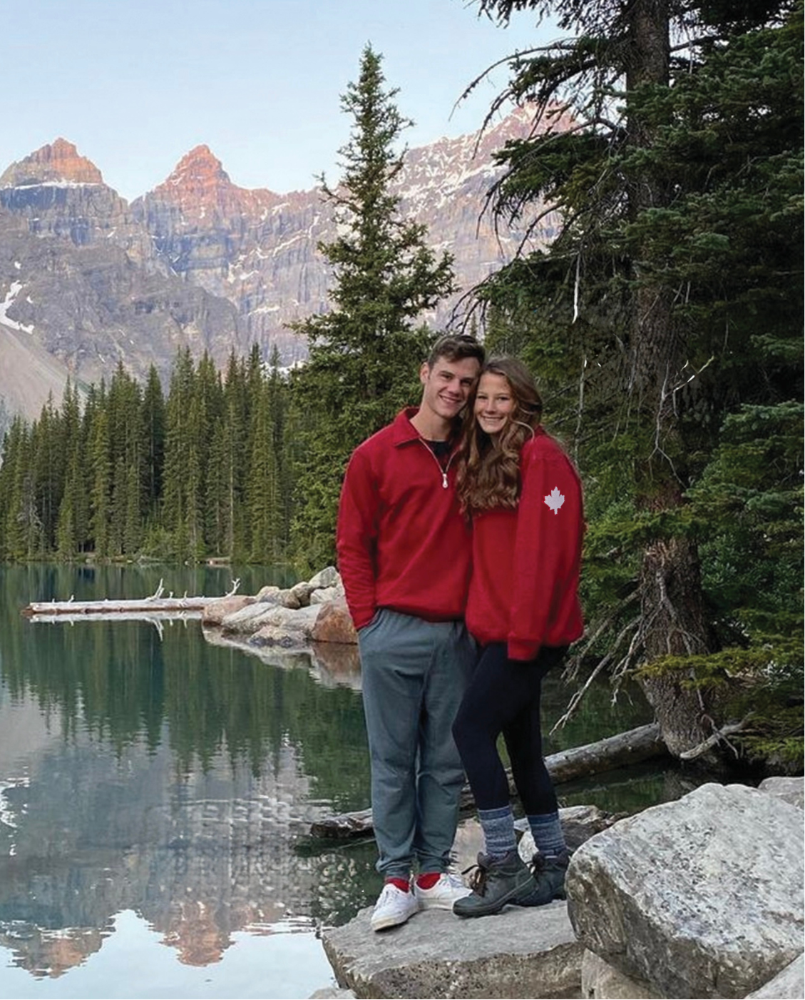 A couple standing in front of Moraine Lake in Banff National Park. Wearing our red canadian jackets with maple leaf patches on the arm. These jackets are made in canada.