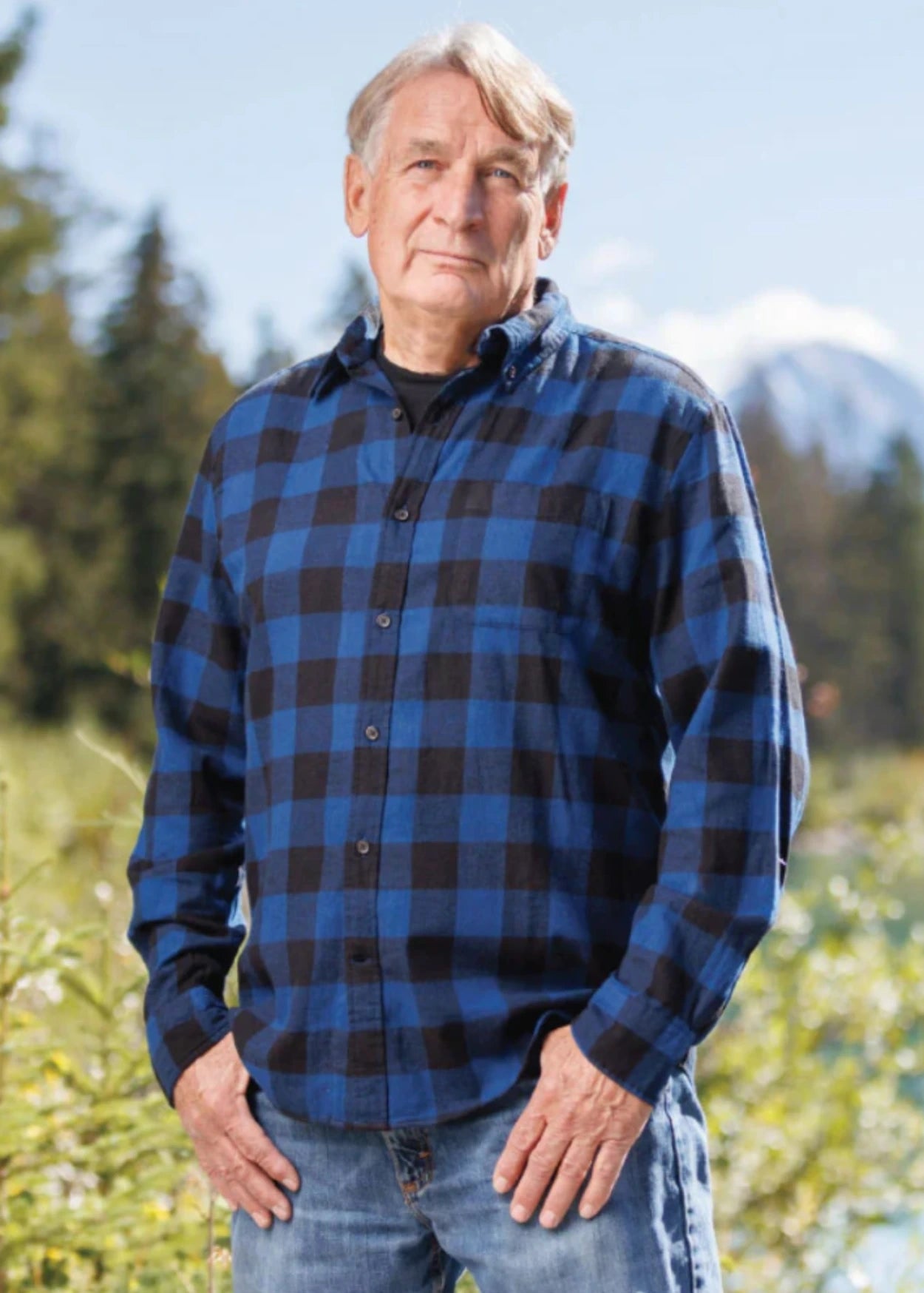 An older man with short hair wearing a long-sleeved marine blue and black buffalo check flannel shirt and blue jeans, standing outdoors.
