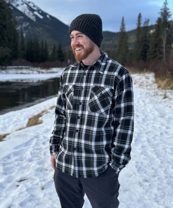 A man wearing a black and white plaid padded flannel jacket, standing near a snowy landscape.