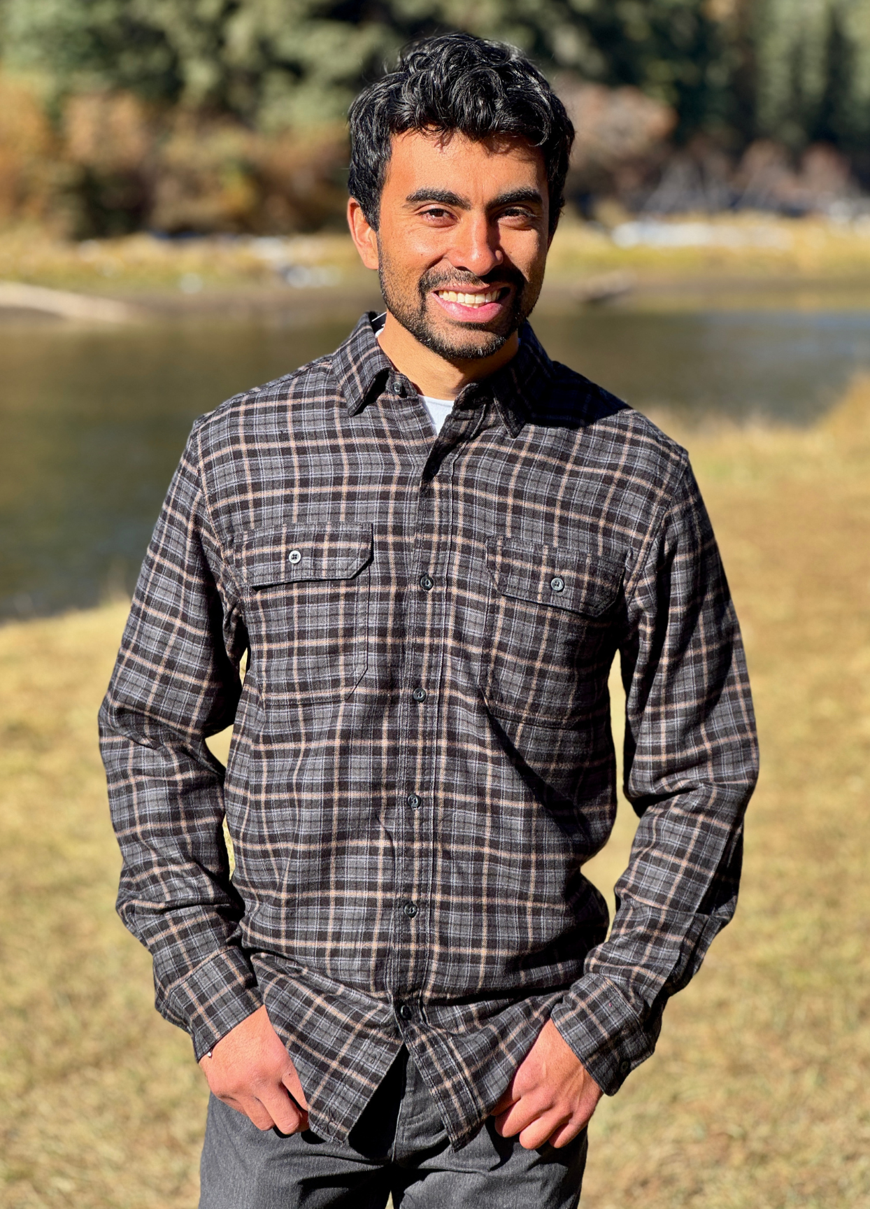 Man wearing a plaid flannel shirt standing outdoors with a river and trees in the background in Banff National Park