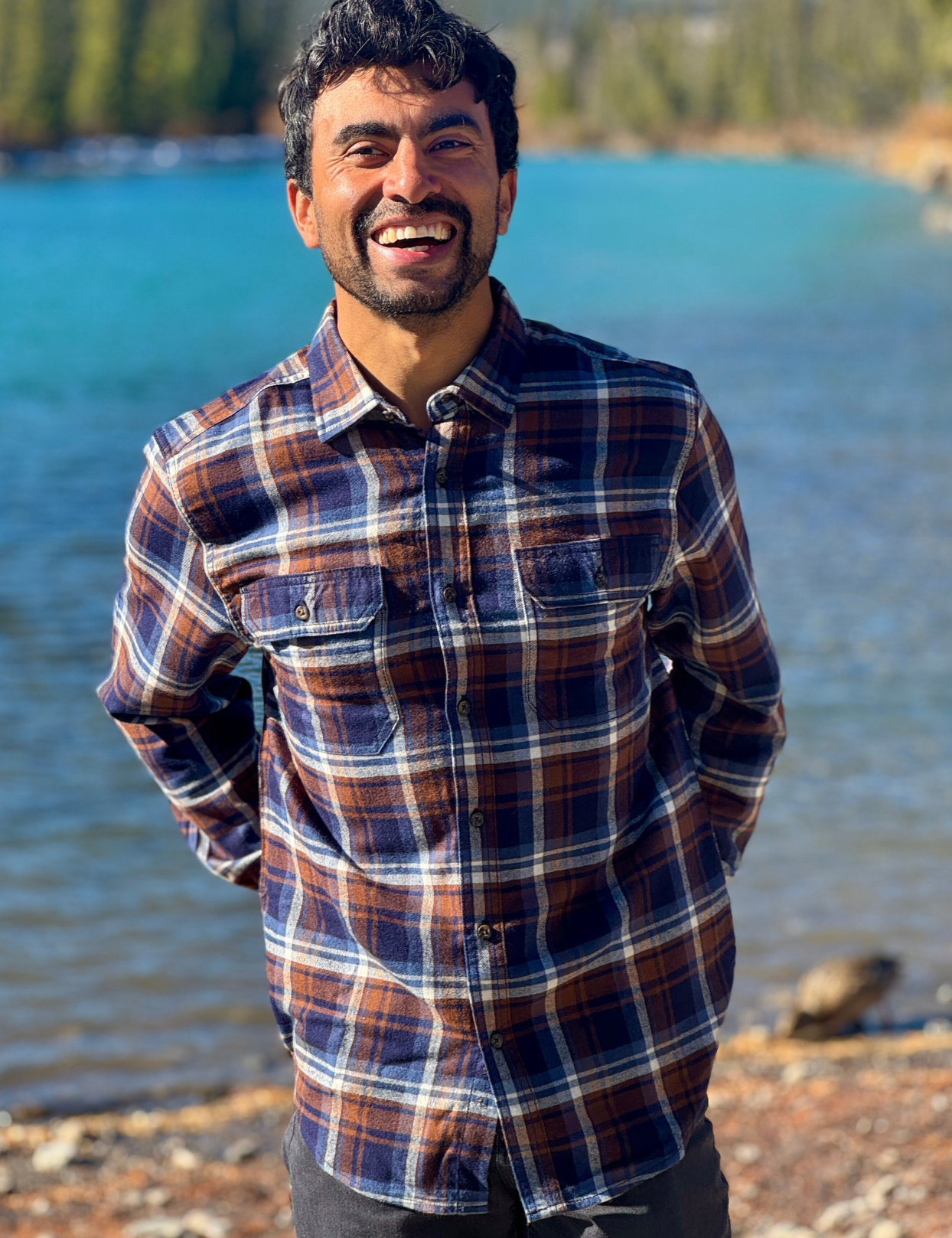 Man wearing a plaid flannel shirt standing by a river with trees in the background in Banff National Park
