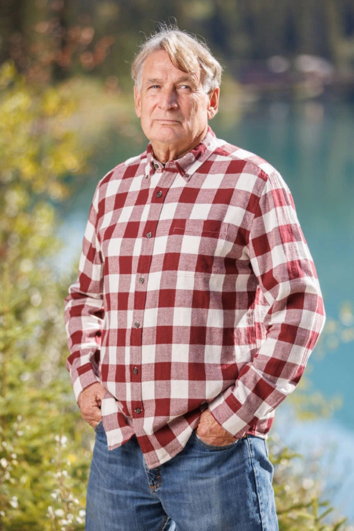 An older man with short grey hair wearing a burgundy and white buffalo check flannel shirt and blue jeans, standing near a body of water.