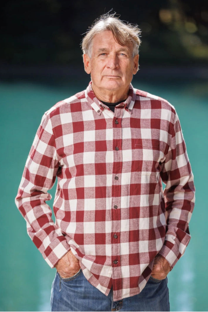 An older man with short grey hair wearing a burgundy and white buffalo check flannel shirt and blue jeans, standing near a body of water.