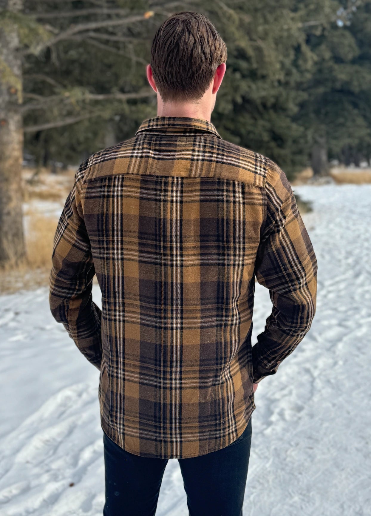 Back view of a man wearing a mocha brown flannel shirt with two flap breast pockets and a shirt tail hemline, standing outdoors.