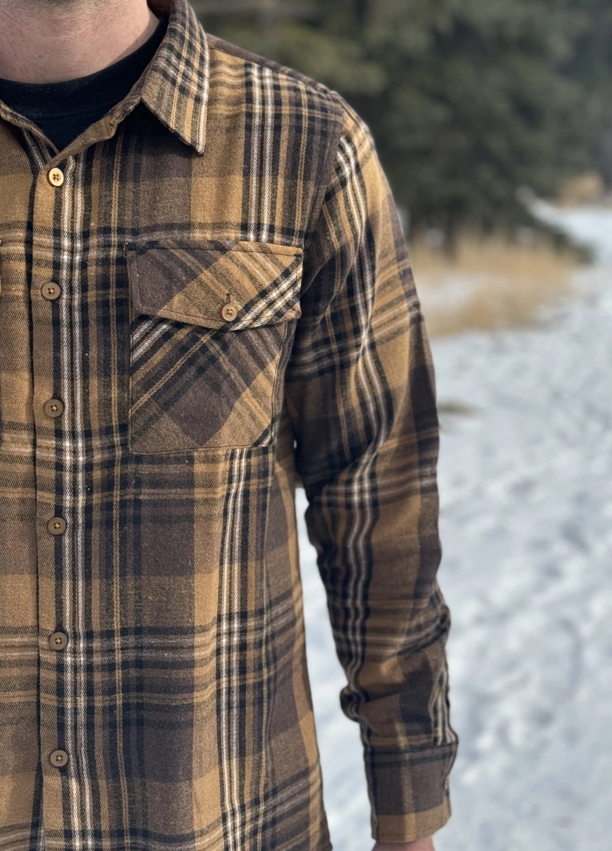 A man wearing a mocha brown flannel shirt with two flap breast pockets and a shirt tail hemline, standing outdoors.