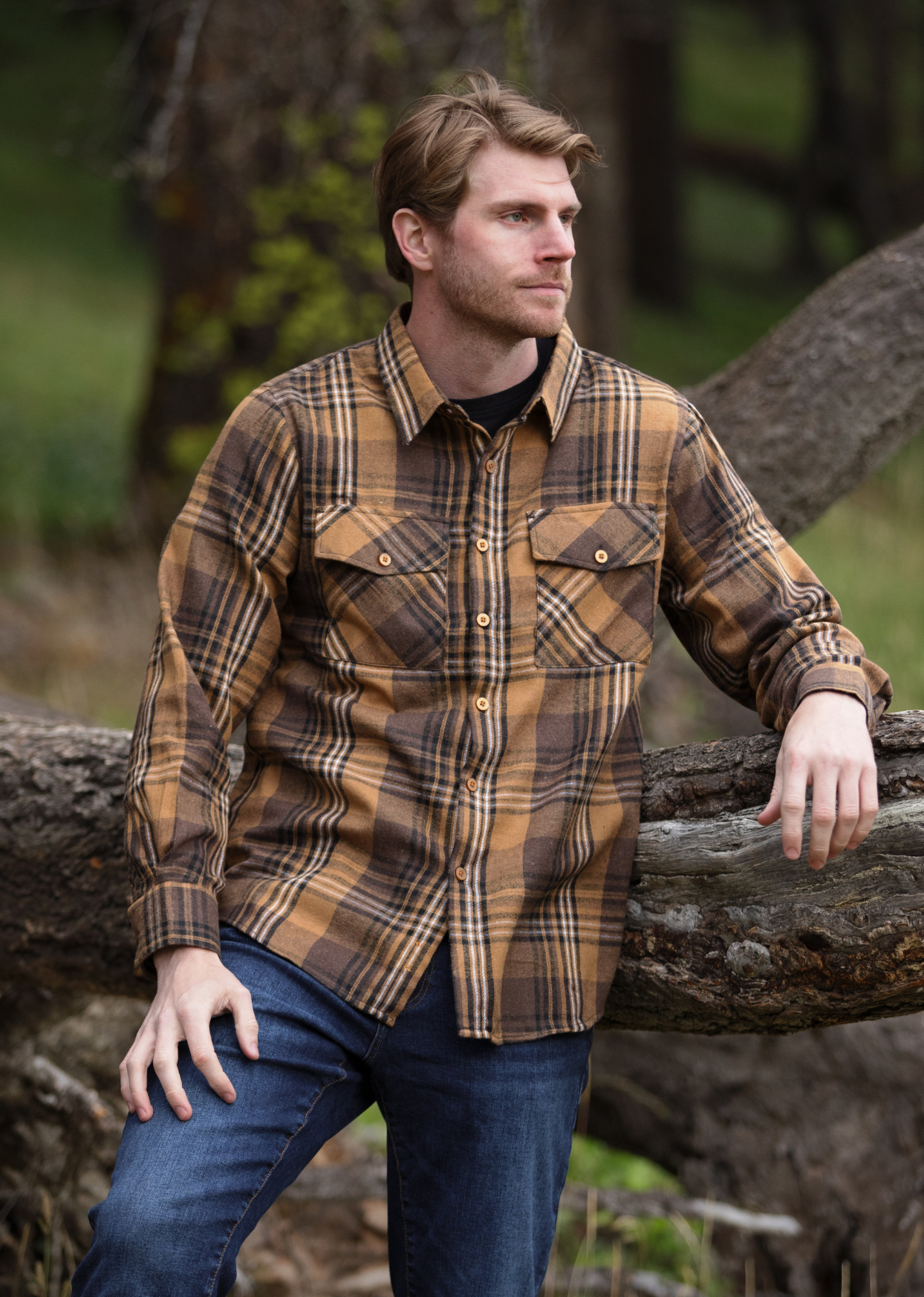 A man wearing a mocha brown plaid flannel shirt with two flap breast pockets and two button cuffs, standing next to a tree.