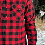 Close up of a man wearing a red and black buffalo check flannel shirt with two flap breast pockets and a button cuff, standing outdoors.