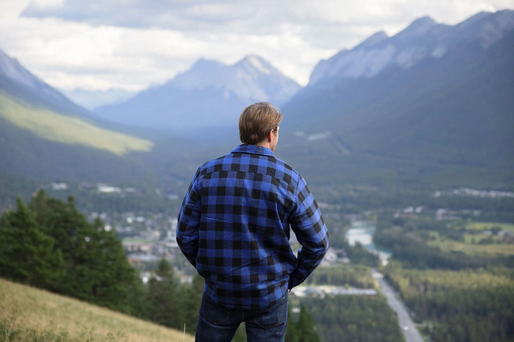 Back view of man wearing a royal blue and black buffalo check padded flannel camping outdoor jacket with jeans. There are mountains in the background