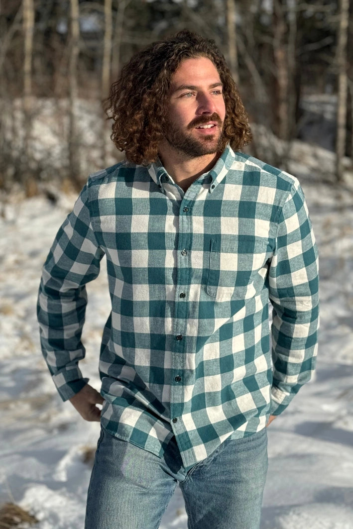 A man standing outdoors wearing a teal buffalo check flannel shirt with long sleeves, button-up collar, and a single breast pocket.