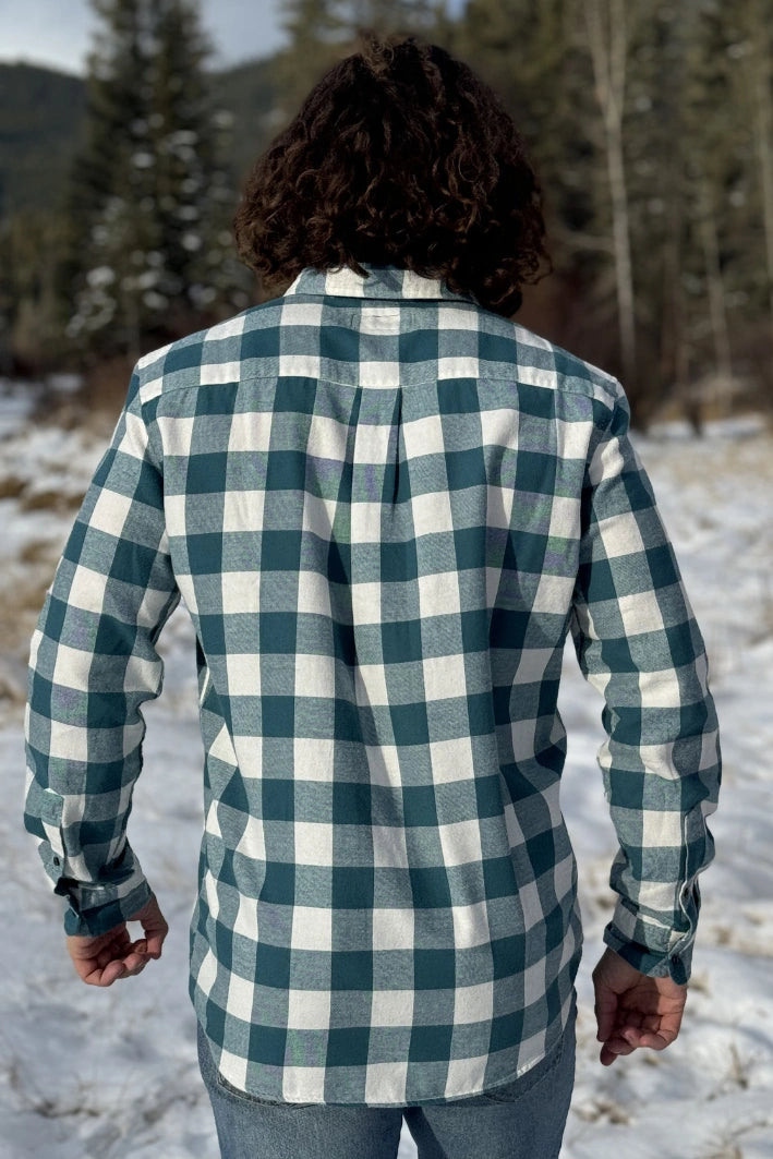 Back view of a man standing outdoors wearing a teal buffalo check flannel shirt with long sleeves, button-up collar, and a single breast pocket.