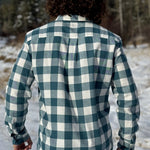 Back view of a man standing outdoors wearing a teal buffalo check flannel shirt with long sleeves, button-up collar, and a single breast pocket.