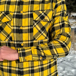 A man wearing a long-sleeved yellow and black flannel plaid shirt with two flap breast pockets and a button cuff.