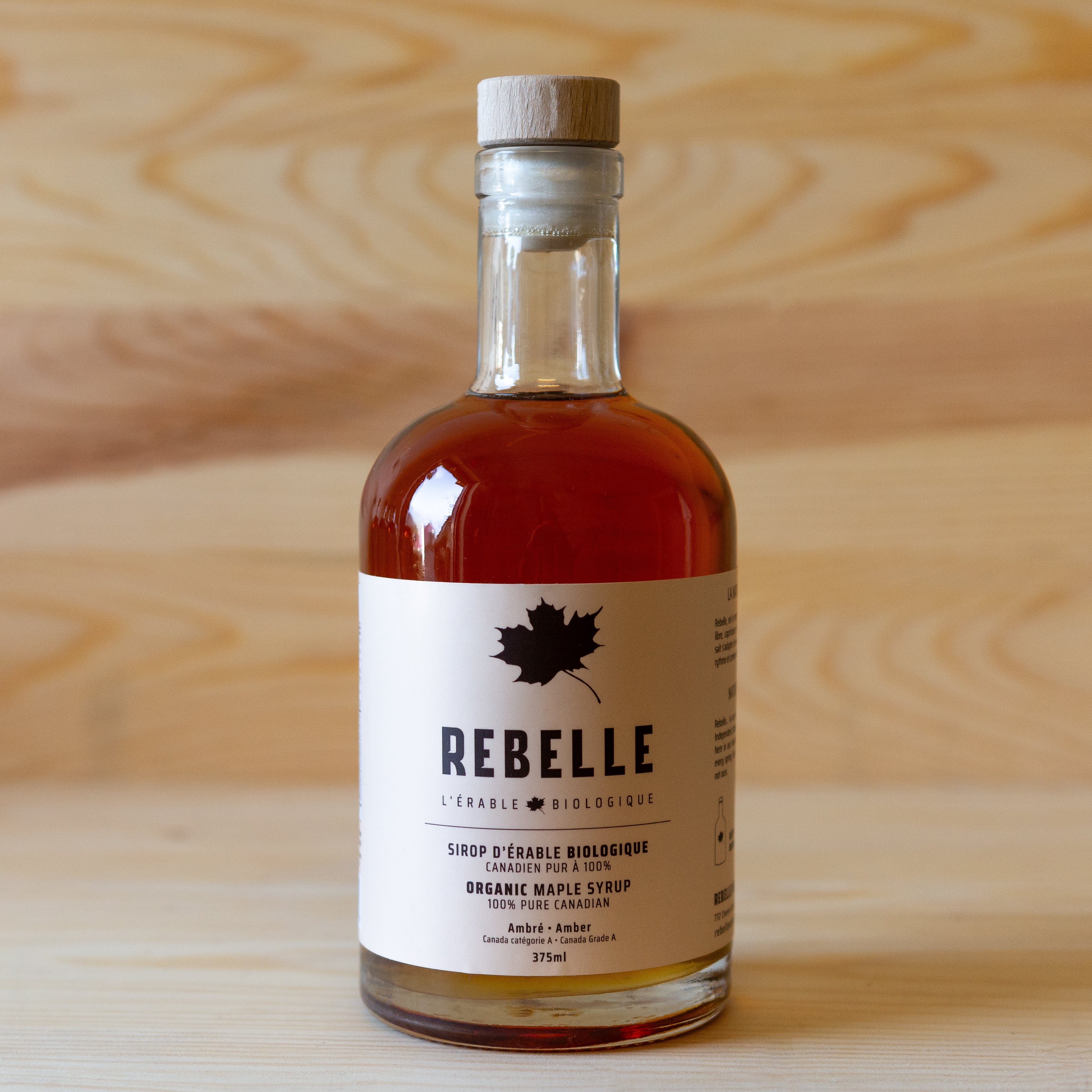 A bottle of Rebelle brand maple syrup with a wooden cap, placed on a wooden surface.