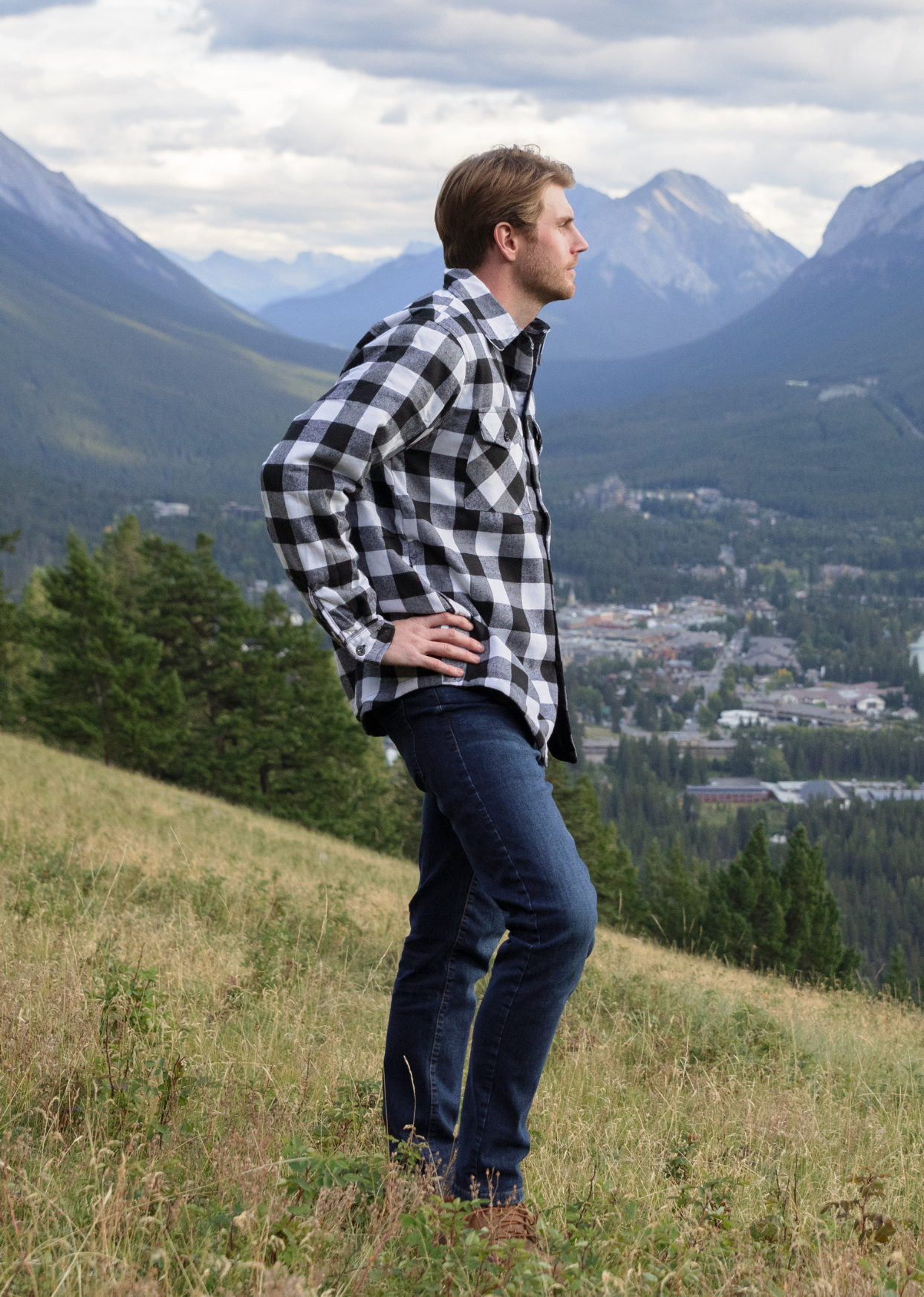 Man standing in front of mountains. He is turned to the side with his hands on his hips. He is wearing our padded flannel camping jacket in black and white buffalo check.