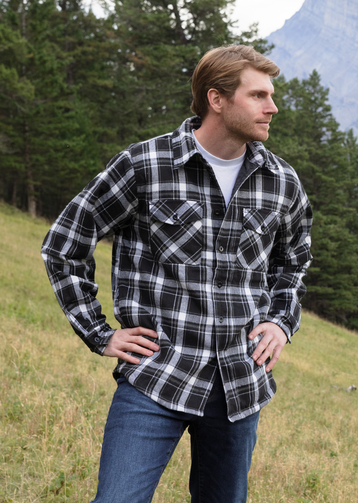 A man wearing a long-sleeved black and white plaid flannel jacket with a shirttail hemline, side pockets, and 2 button cuffs, standing outdoors.