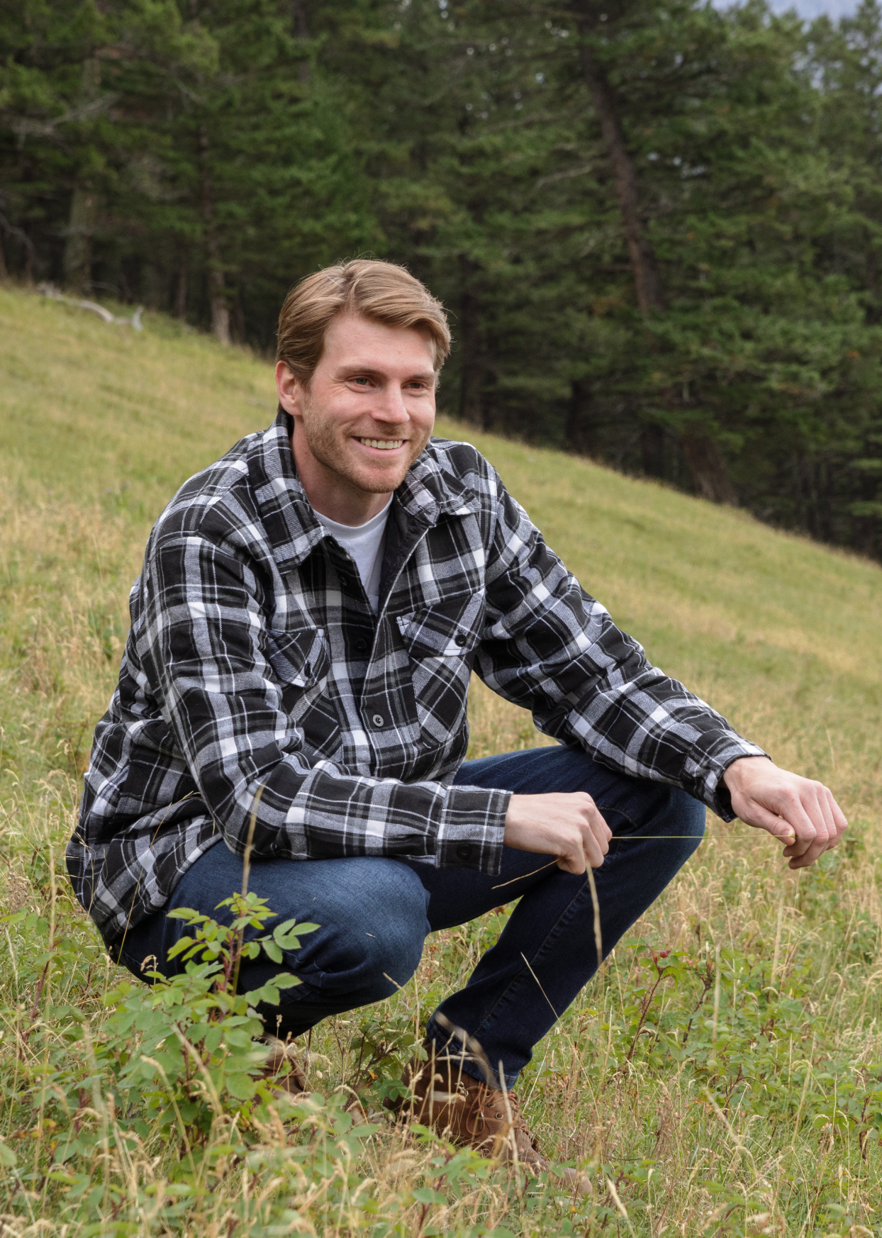 A man wearing a long-sleeved black and white plaid flannel jacket with a shirttail hemline, side pockets, and 2 button cuffs, sitting outdoors.