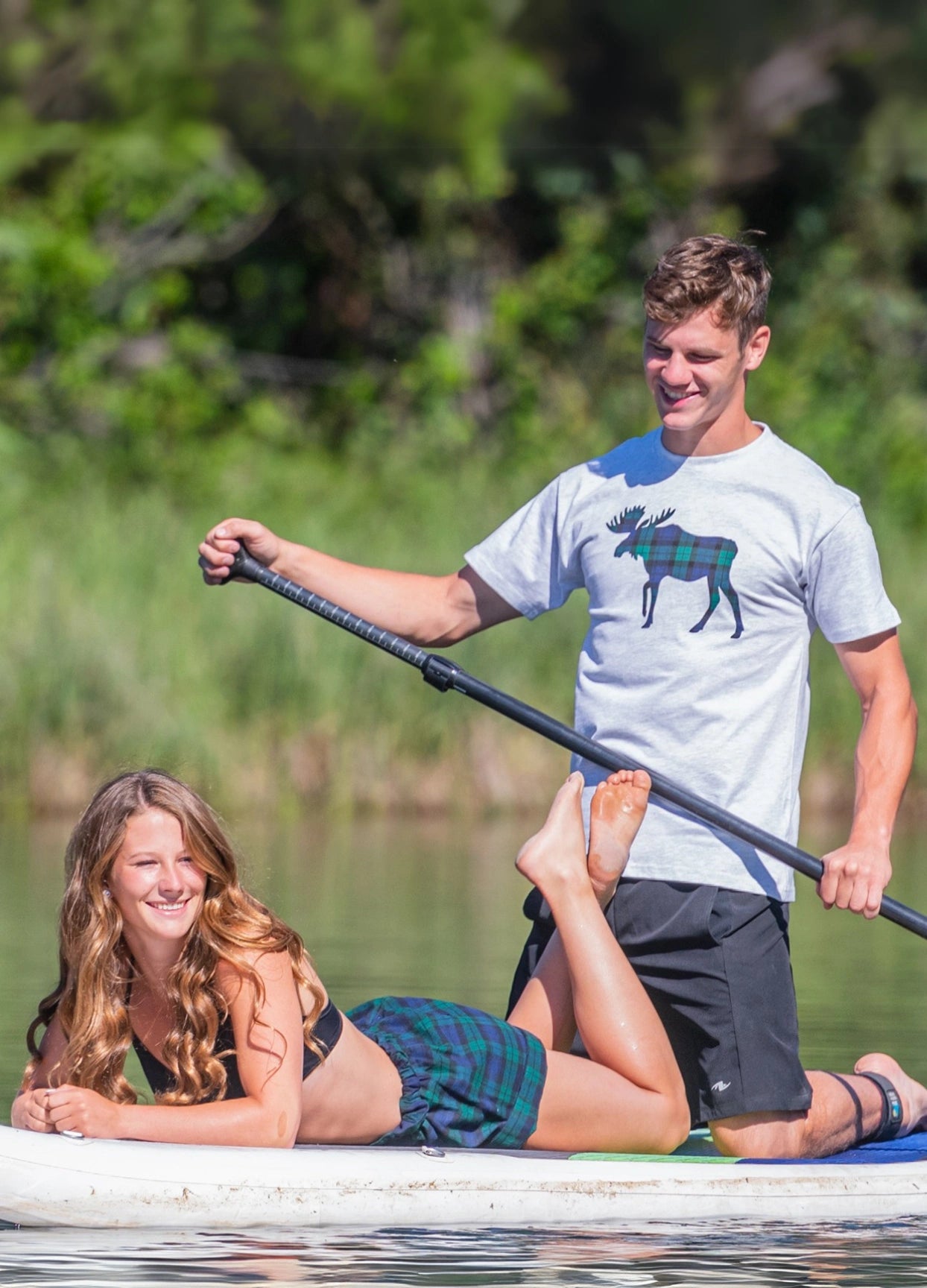 A woman sitting on a paddleboard wearing black watch shorts and a man standing next to her with a paddle, both smiling.