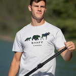 Man wearing a light grey t-shirt with Banff Canada design, holding a paddle by a body of water.