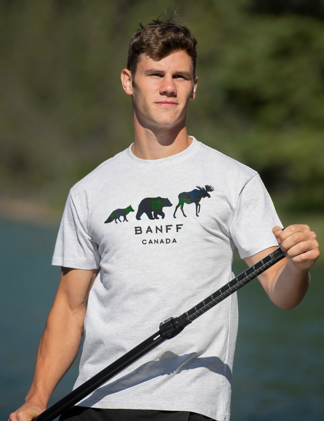 Man wearing a white t-shirt with Banff Canada design, holding a paddle by a body of water.