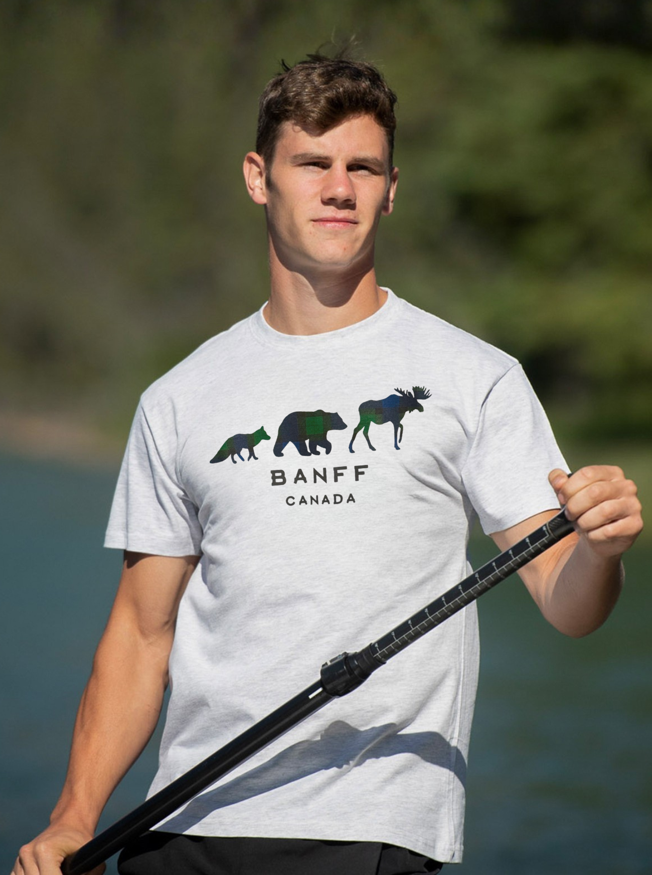 Man wearing a light grey t-shirt with Banff Canada design, holding a paddle by a body of water.