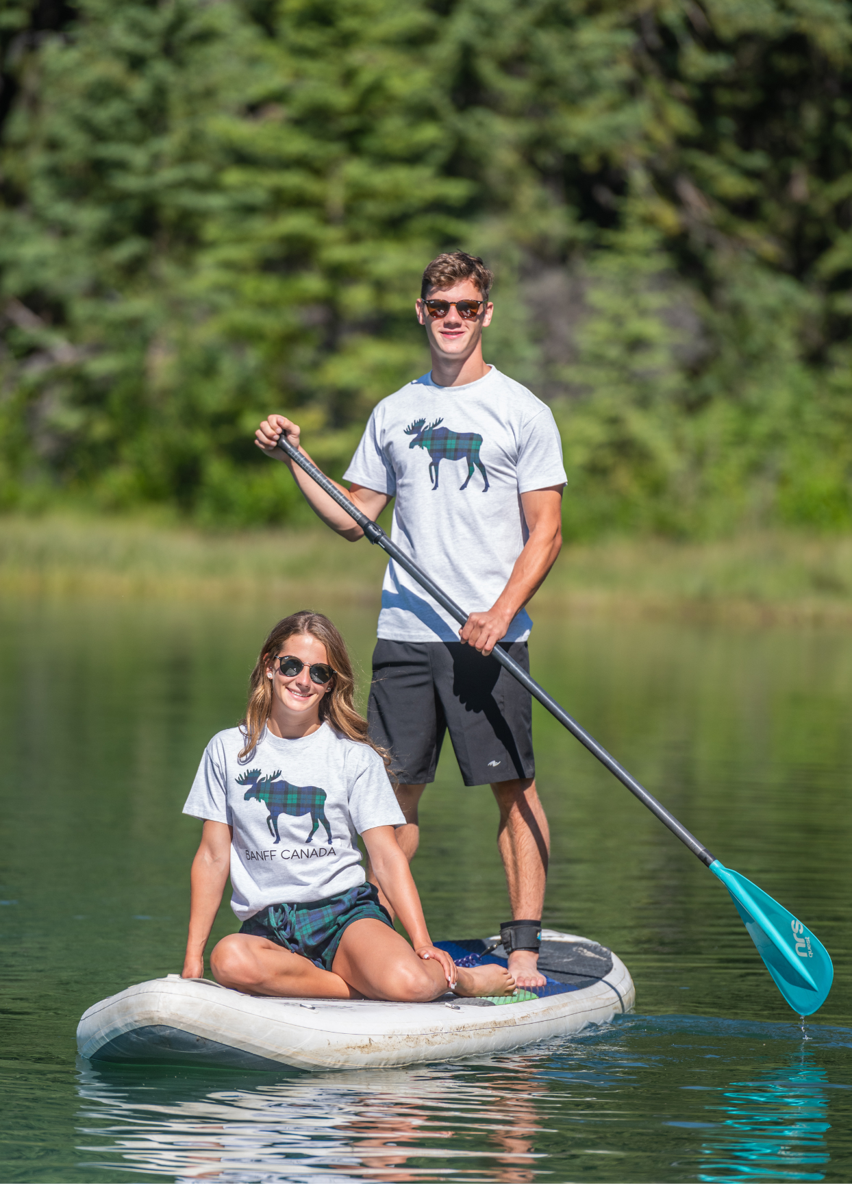 Two people paddle boarding on a lake with trees in the background. Wearing our grey and black watch Canadian animals t-shirts