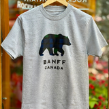 Grey t-shirt with a bear graphic and 'Banff Canada' text on a hanger against a blurred background.