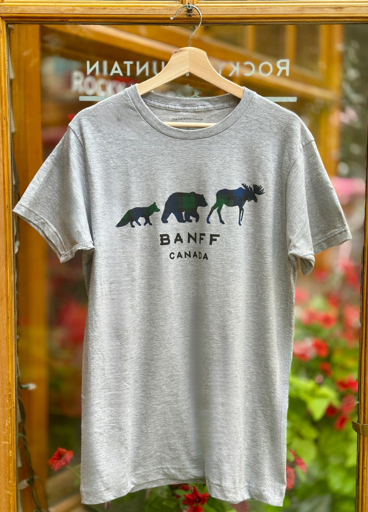 Grey t-shirt with bear and moose graphic and 'Banff Canada' text on a hanger against a blurred floral background.