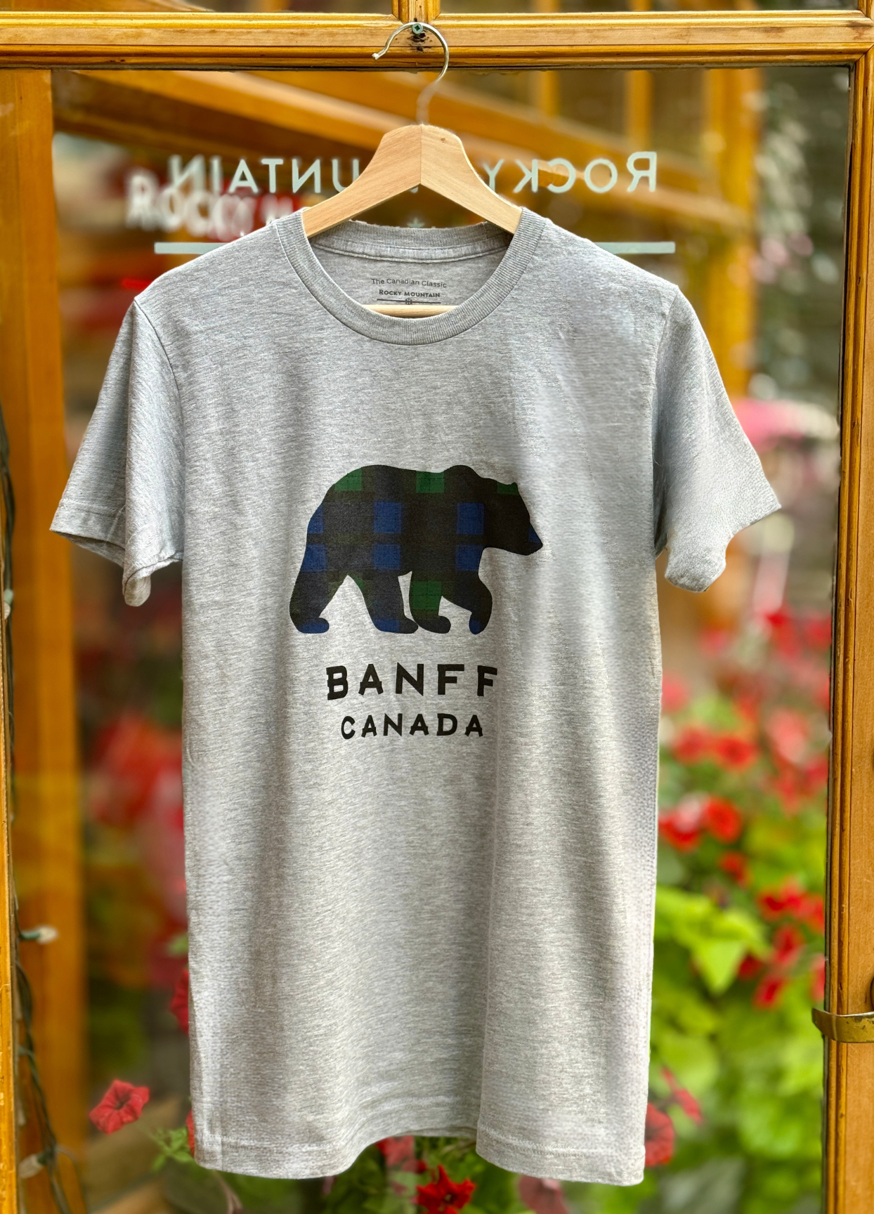 Gray t-shirt with a bear graphic and 'Banff Canada' text on a hanger in front of a store window.