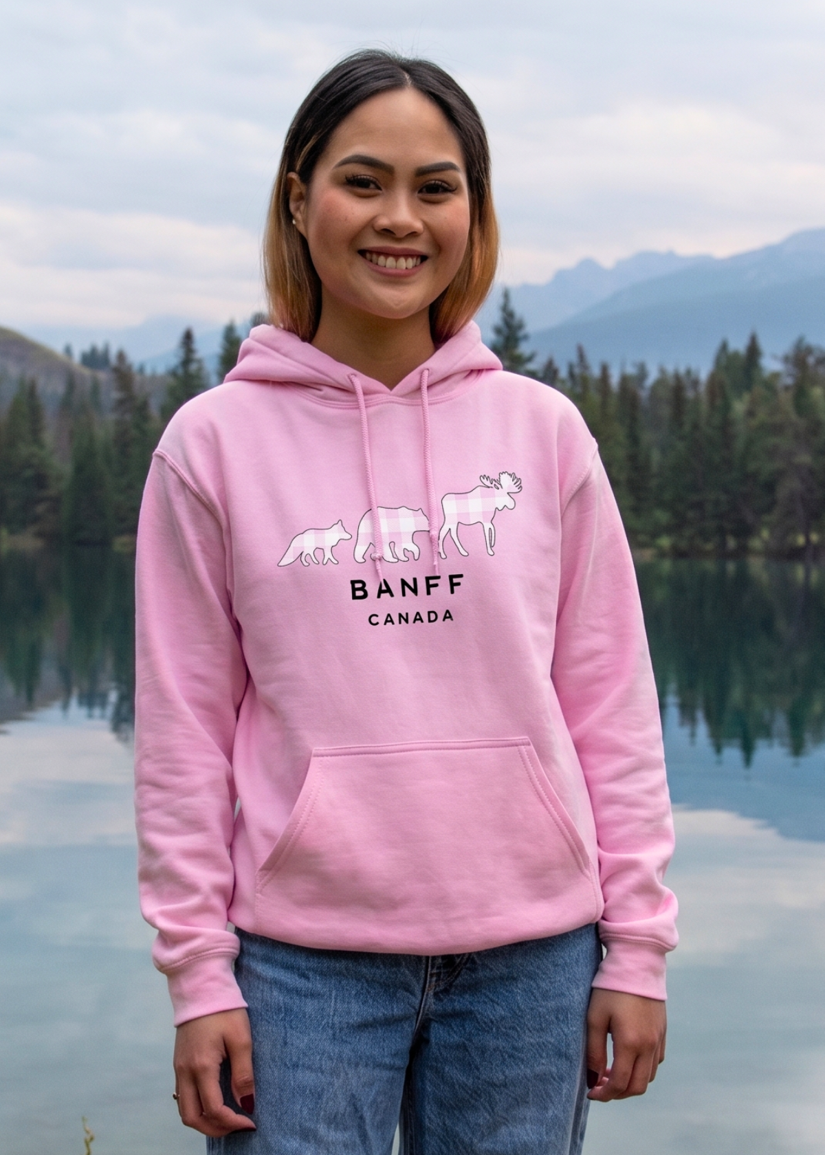 Person wearing a pink hoodie with 'Banff Canada' design by a lake and mountains.