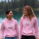 Two people wearing pink Banff hoodies by a lake with trees in the background
