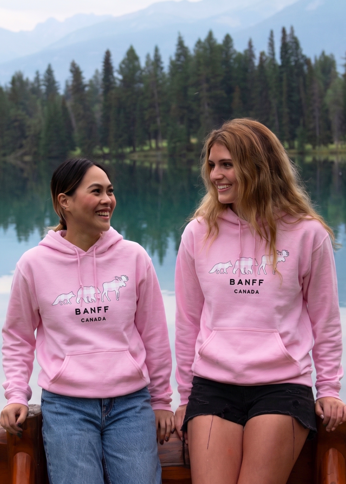 Two people wearing pink Banff hoodies by a lake with trees in the background