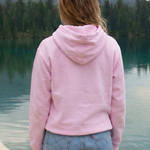 Person wearing a pink hoodie and denim shorts standing by a lake with mountains in the background